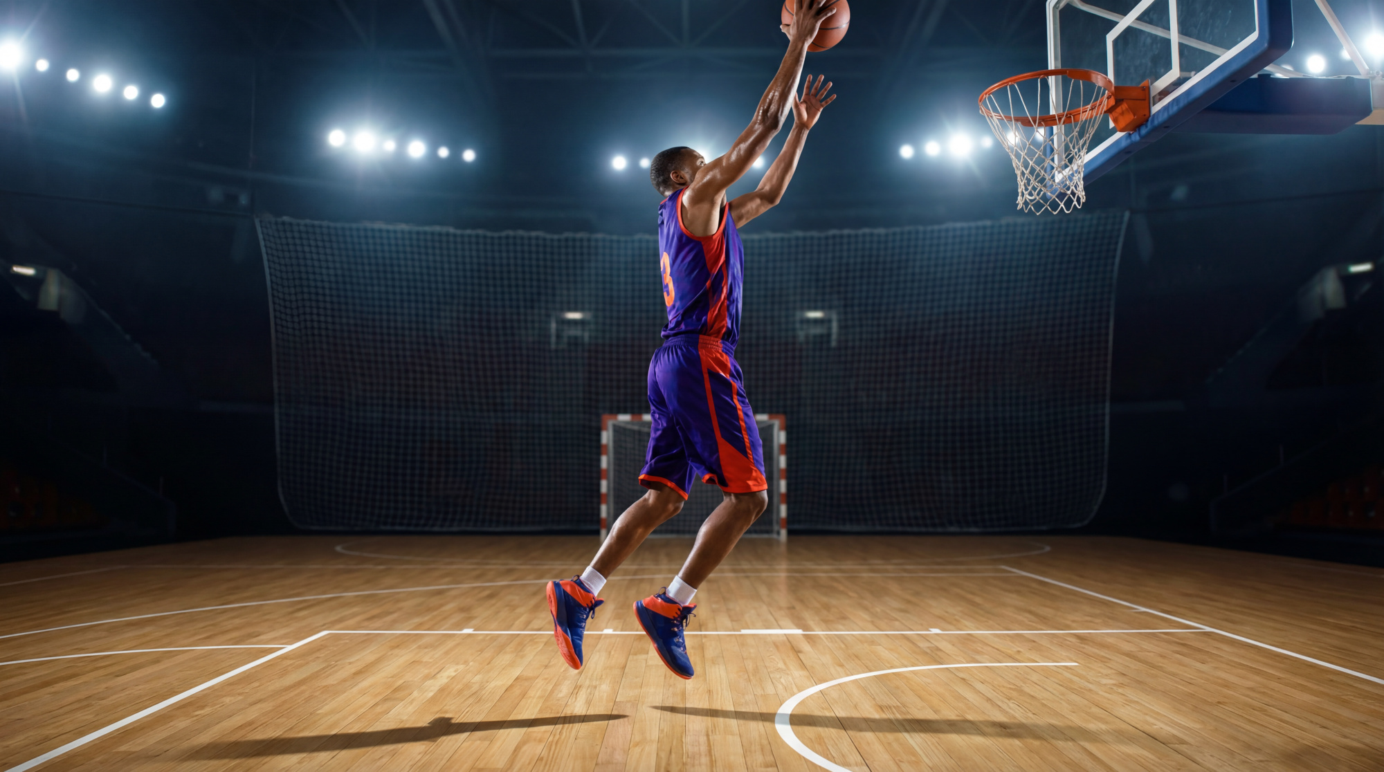 Joueur de basket athlétique en plein saut, portant un maillot violet et orange, sur un terrain en bois éclairé, visant le panier.